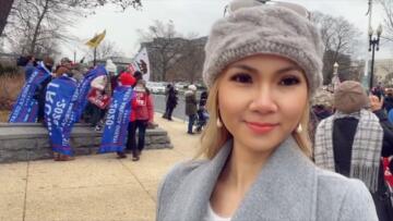 Screenshot of Sonia Ohlala looking to the side of the camera, wearing a gray coat and beanie; behind her is a group of protestors wearing Trump 2020 MAGA flags wrapped around them